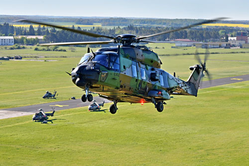 Depuis un NH90 Caïman au décollage, vue sur un second appareil quittant simultanément la base de Chaumont, avec au sol d’autres hélicoptères alignés le long de la piste