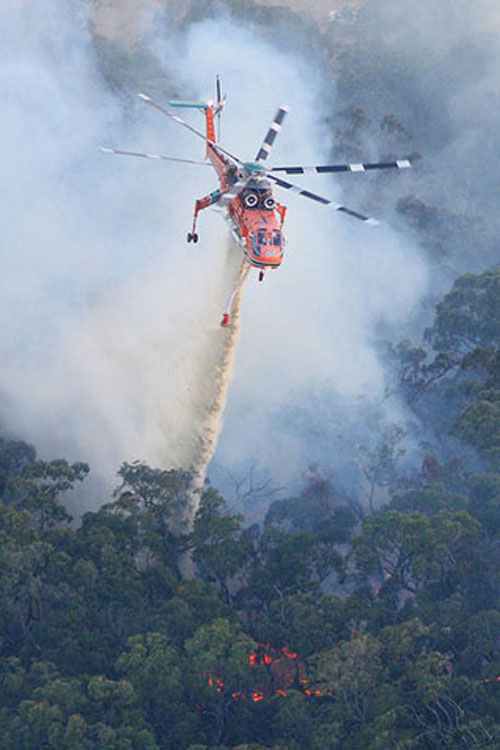 Un hélicoptère S-64 Air Crane largue sa charge d'eau au-dessus des flammes d'un incendie de forêt en montagne
